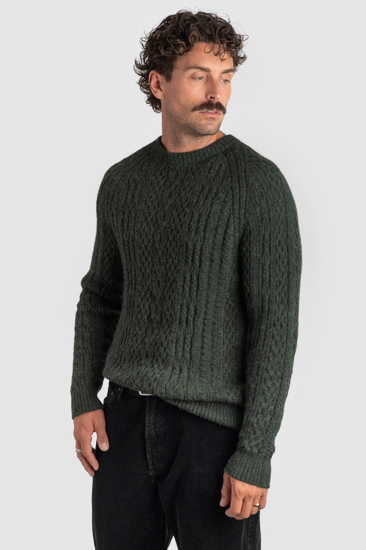 Man wearing a dark green cable knit sweater against a light gray background #colour_pine