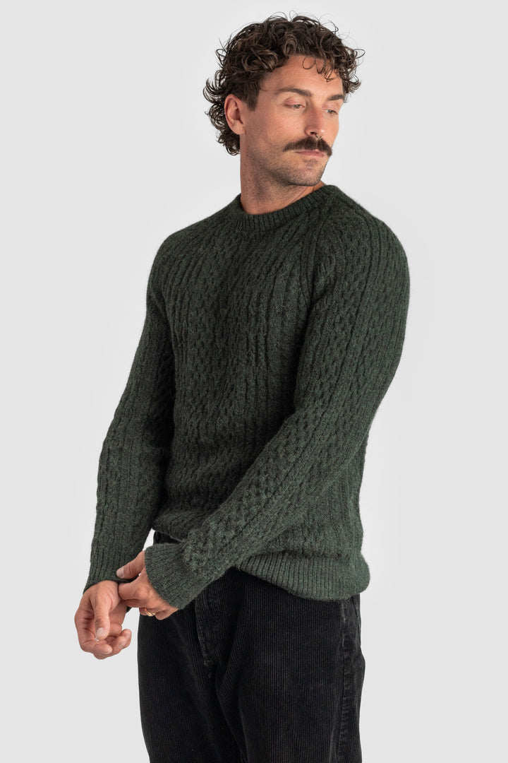 Man wearing a dark green cable knit sweater against a light gray background #colour_pine