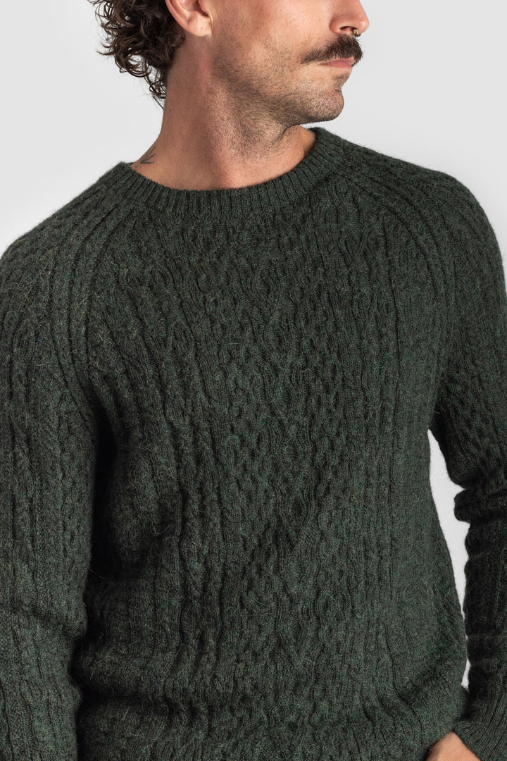 Man wearing a dark green cable knit sweater against a plain background #colour_pine